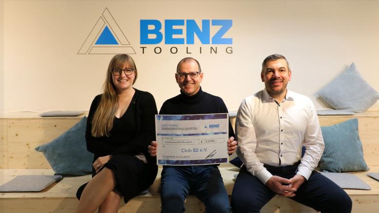 Mitarbeiter von  BENZ und Club 82 sitzen vor einem BENZ Logo und zeigen einen Spendenscheck