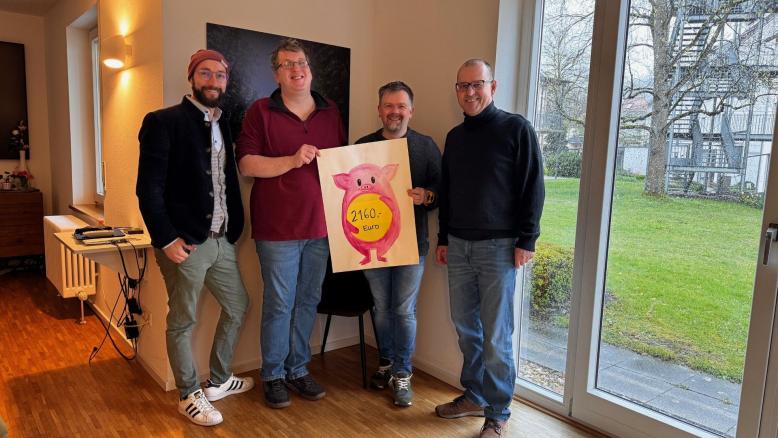 4 Personen stehen vor einer Wand und halten ein Spendenschild in der Hand. 